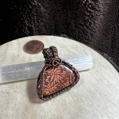 One-of-a-kind Starburst Jasper pendant wrapped in reclaimed copper wire displayed on a selenite wand with penny for size reference.