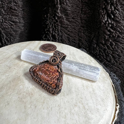One-of-a-kind Starburst Jasper pendant wrapped in reclaimed copper wire displayed on a selenite wand with penny for size reference.