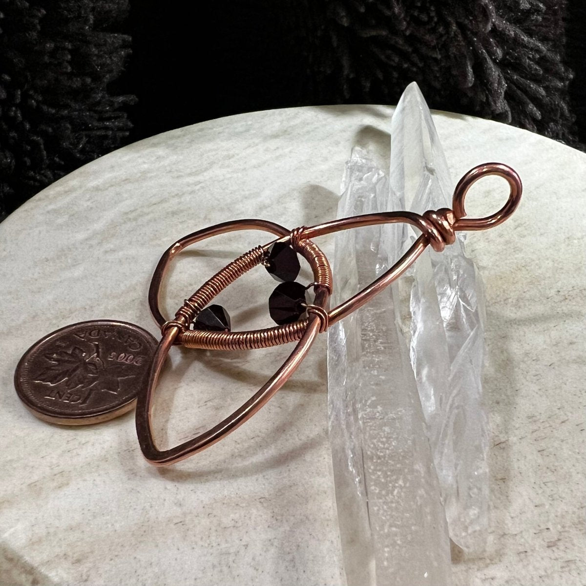 Copper wirependant with garnet beads on a marble surface next to a penny and crystal.