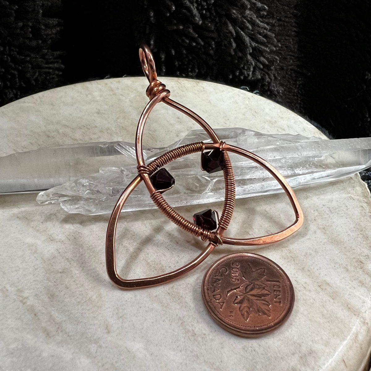 Copper wire-wrapped pendant with garnet stones on a marble surface with a penny for scale.