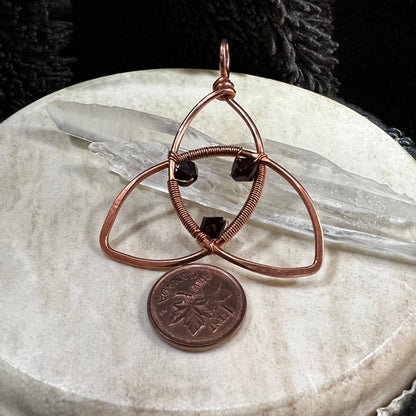 Copper pendant with three interlocking triangles and a penny on a beige surface leaning on quartz crystals