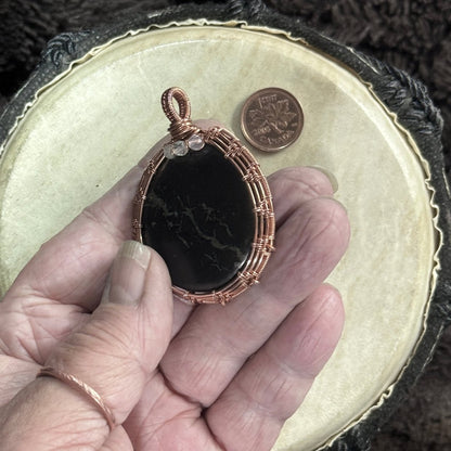 Hand-held front view of a tactile polished shungite pendant with a river-like pyrite seam, pale faceted fluorite and bright raw copper weaving.