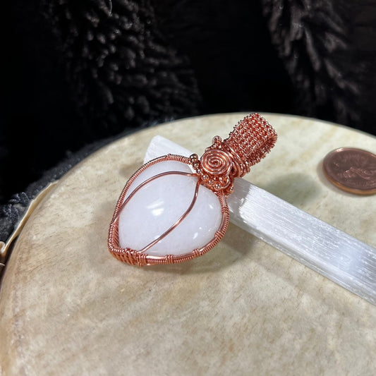 Copper-wrapped white quartz crystal heart pendant on a beige surface with a coin for scale