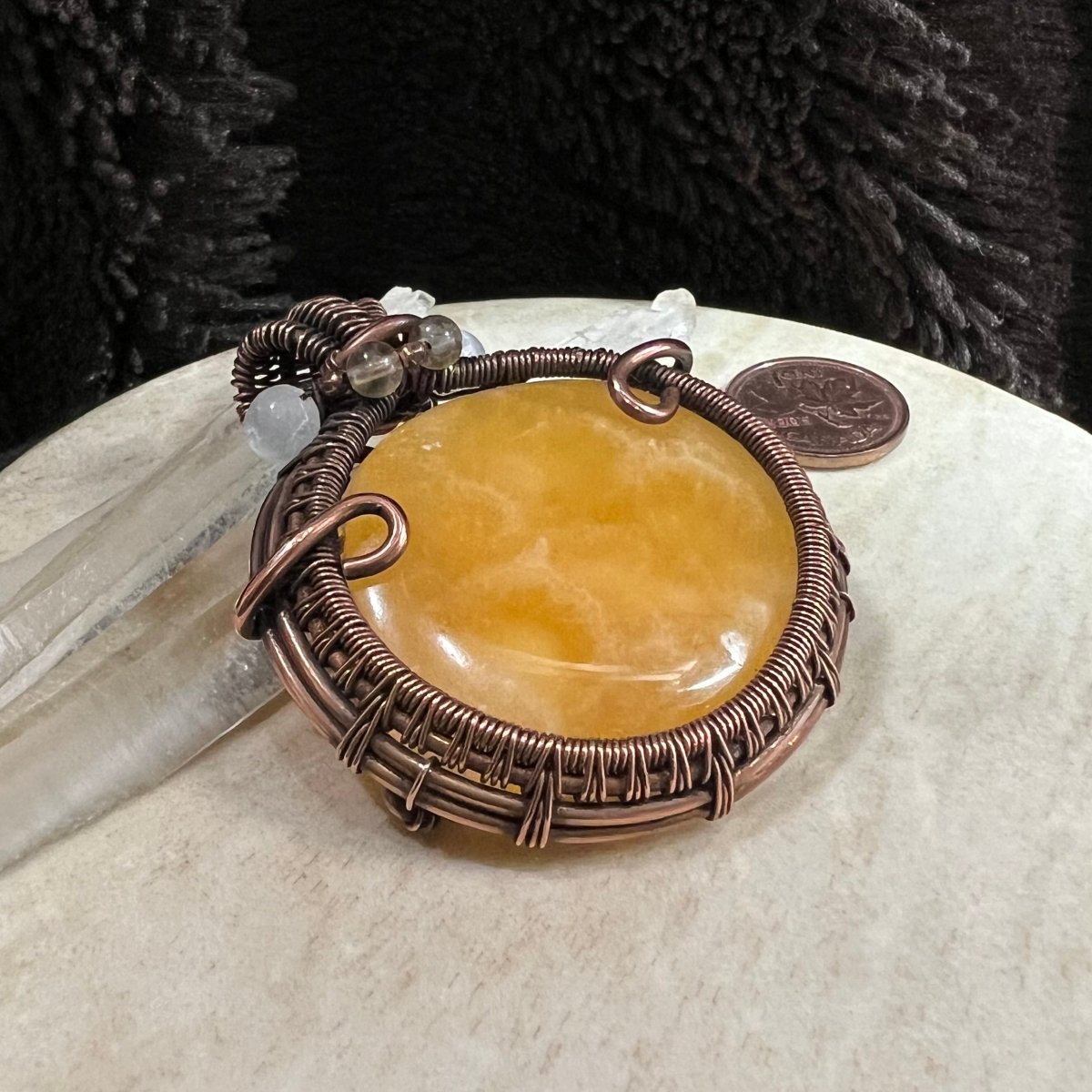 Close-up of yellow calcite stone in copper wire wrap showing intricate weaving and polished surface glow, next to CAnadian penny for scale